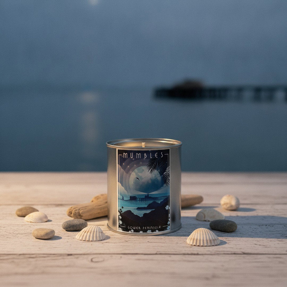 Mumbles Gower Peninsula scented candle in a silver tin on driftwood decking with seashells, sea and pier at dusk behind